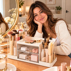 A woman with long brown hair rests her head on her hand, smiling at a vanity table. In front of her is a white and clear acrylic makeup organizer filled with cosmetics, brushes, and skincare items.